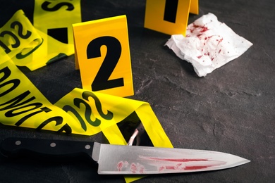 Bloody knife, yellow tape and evidence marker on black slate table, closeup. Crime scene