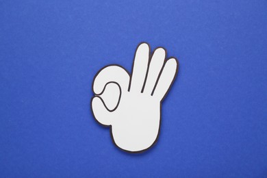 Photo of Paper cutout of okay hand gesture on blue background, top view