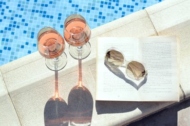 Photo of Glasses of tasty rose wine, open book and sunglasses on swimming pool edge