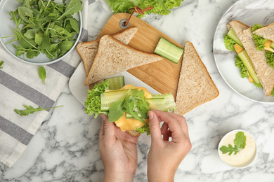 Woman adding arugula to tasty sandwich at white marble table, top view
