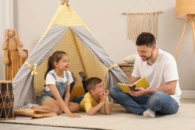Father reading book to children near toy wigwam at home