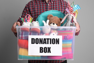 Man holding donation box full of different toys, clothes and stationery on grey background, closeup