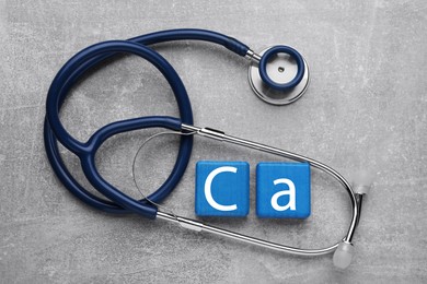Photo of Light blue cubes with symbol Ca (Calcium) and stethoscope on gray background, top view