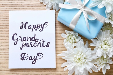Beautiful white flowers, gift box and card with phrase Happy Grandparents Day on wooden background, flat lay