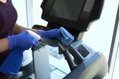 Woman cleaning treadmill with disinfectant spray and cloth in gym, closeup