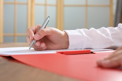 Woman writing on sheet of paper in red folder at wooden table in office, closeup