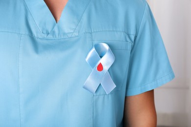 Doctor with light blue ribbon and paper blood drop indoors, closeup. World Diabetes Day