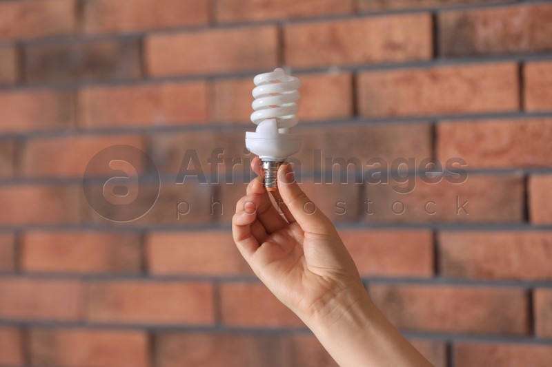 Woman holding fluorescent light bulb near brick wall indoors, closeup. Saving energy concept