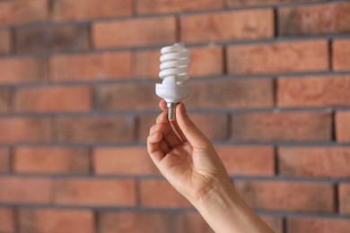 Woman holding fluorescent light bulb near brick wall indoors, closeup. Saving energy concept
