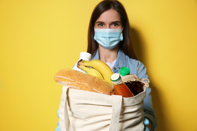 Female volunteer in protective mask and gloves with products against yellow background, focus on bag. Aid during coronavirus quarantine