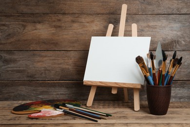 Easel with blank canvas, palette and brushes on wooden table