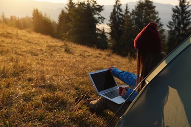 Woman working on laptop near camping tent outdoors surrounded by beautiful nature