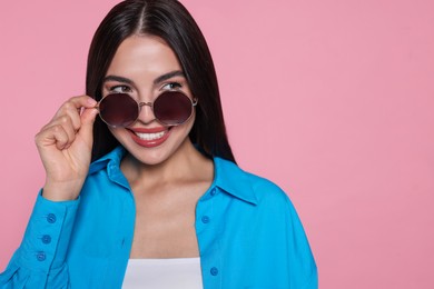 Attractive happy woman touching fashionable sunglasses against pink background, closeup. Space for text