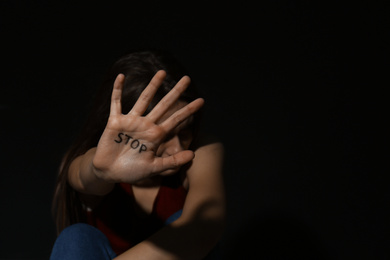 Photo of Abused young woman showing palm with word STOP against black background, focus on hand and space for text. Domestic violence concept