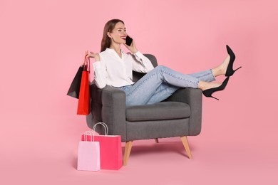 Photo of Happy young woman with shopping bags and smartphone on armchair against light pink background. Big sale
