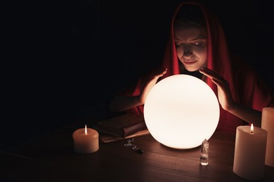 Soothsayer using glowing crystal ball to predict future at table in darkness, space for text. Fortune telling