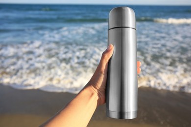 Photo of Woman holding metallic thermos with hot drink on beach near sea, closeup