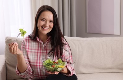 Beautiful overweight woman eating salad in living room, space for text. Healthy diet
