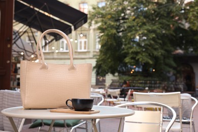 Stylish bag and cup of coffee on white table in outdoor cafe, space for text