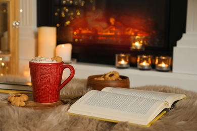 Cup of cocoa, book and cookies near fireplace indoors