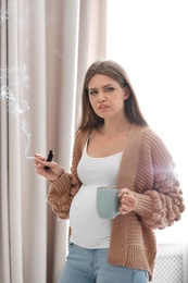 Young pregnant woman with cup of coffee smoking cigarette at home. Harm to unborn baby