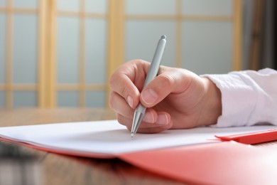 Woman writing on sheet of paper in red folder at wooden table in office, closeup