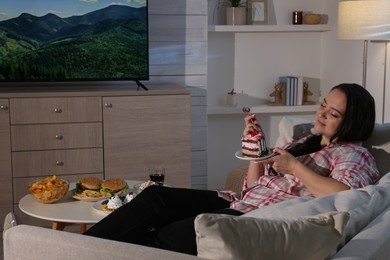 Happy overweight woman eating cake while watching TV on sofa at home