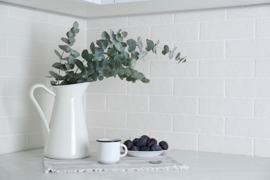 Photo of Beautiful eucalyptus branches, cup of drink and fresh plums on countertop in kitchen, space for text. Interior element