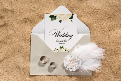 Envelope with wedding invitation, brooch and gold rings on sandy beach, top view