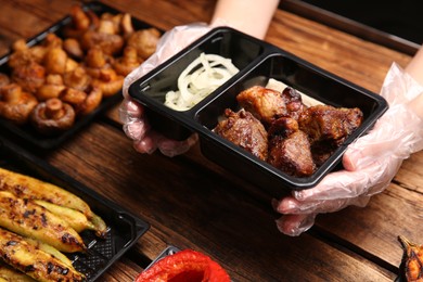 Waiter with plastic container of tasty shish kebab at wooden table, closeup. Food delivery service