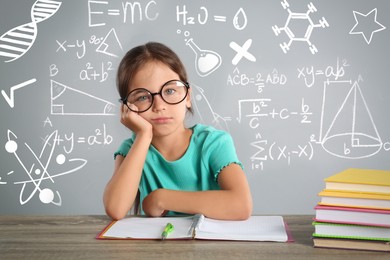 Bored little girl doing homework at wooden table and different formulas against light grey background