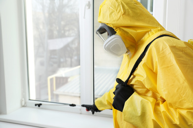 Pest control worker in protective suit spraying insecticide on window sill indoors