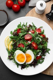 Photo of Delicious salad with boiled egg, arugula and tomatoes served on black table, flat lay