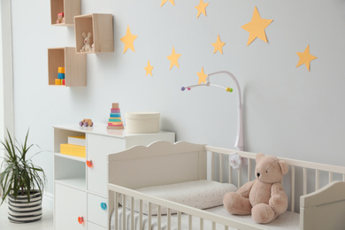Crib with toy bear and mobile in stylish baby room interior