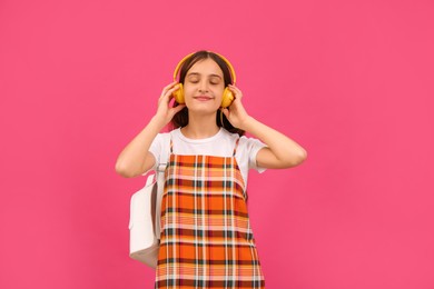 Teenage student with backpack and headphones listening to music on pink background