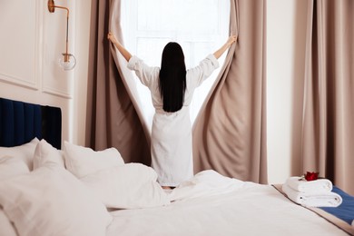 Young woman opening window curtains in hotel room, back view