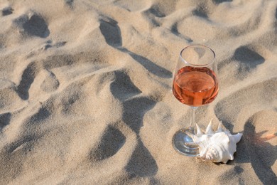 Photo of Glass of tasty rose wine and seashell on sand, space for text