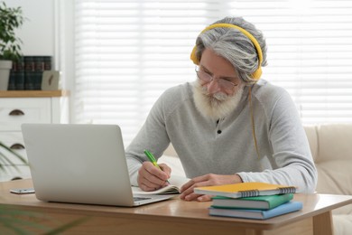 Middle aged man with laptop, notebook and headphones learning at table indoors