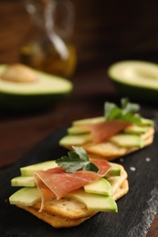 Photo of Delicious crackers with avocado, prosciutto and parsley on slate board, closeup