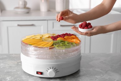 Woman putting raspberry into fruit dehydrator machine at grey marble table in kitchen, closeup