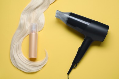 Photo of Spray bottle with thermal protection, hairdryer and lock of blonde hair on yellow background, flat lay