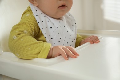 Cute little baby wearing bib in highchair indoors, closeup