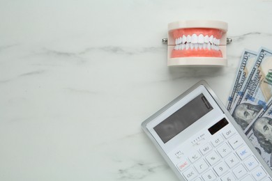 Educational dental typodont model, dollar banknotes and calculator on white table, flat lay with space for text. Expensive treatment