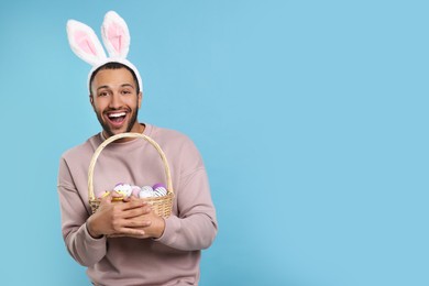 Photo of Happy African American man in bunny ears headband holding wicker basket with Easter eggs on light blue background, space for text