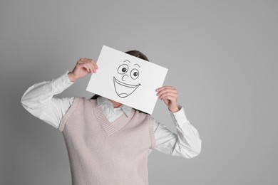 Photo of Woman hiding behind sheet of paper with happy face on grey background, space for text