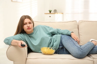 Lazy young woman with bowl of chips watching TV on sofa at home