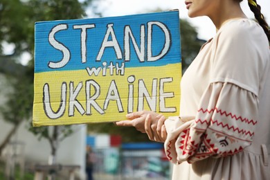 Young woman holding poster in colors of national flag and words Stand with Ukraine outdoors, closeup