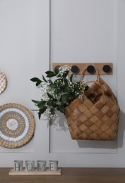 Stylish wicker basket with bouquet of flowers hanging on wooden rack indoors
