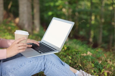 Young woman with cup of coffee working on laptop in forest, closeup
