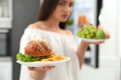 Concept of choice. Woman holding fruits and burger with French fries in kitchen, closeup
